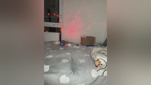A video of a cluttered bedroom with a white bedspread adorned with cloud and duck patterns. A glowing red light casts a warm hue, creating a cozy atmosphere. Toys and a small box are scattered on the floor.