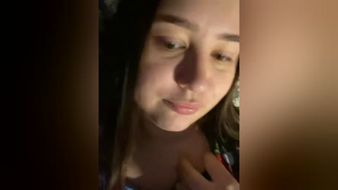 A close-up video of a young woman with light skin and straight, dark hair. She has a neutral expression, wearing light makeup, and a soft, warm light illuminates her face.