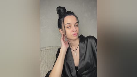 Video of a light-skinned woman with dark hair in a top knot, wearing a black satin robe, and a thin necklace. She's indoors, leaning against a beige wall with a textured, light-colored fabric behind her.
