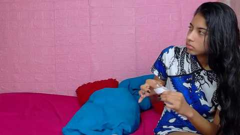 Video of a young woman with long black hair in a blue and white patterned dress, sitting on a pink bed with red and blue pillows, looking thoughtfully at a white object.