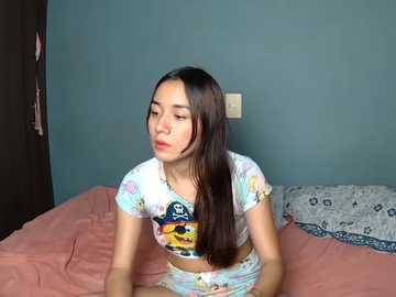 Video of a young Asian woman with long dark hair, wearing a colorful tie-dye crop top and matching shorts, sitting on a pink bedspread against a teal wall.