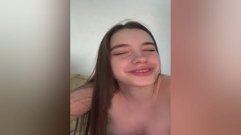 A video of a smiling young woman with straight brown hair, fair skin, and closed eyes, partially nude, against a blurred beige and pink background.