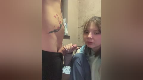 Video of a naked man with a tattoo holding his erect penis in front of a young girl with long hair, wearing a blue hoodie, in a bathroom with a beige wall.