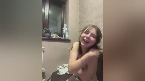 Video of a topless, laughing young woman with light skin and long blonde hair, sitting in a cramped, dimly lit bathroom with beige walls and a window.