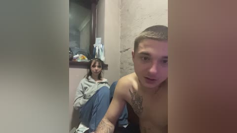 Video of a young, shirtless man with tattoos and a woman in a gray hoodie and jeans sitting on a toilet in a small, dimly-lit bathroom.