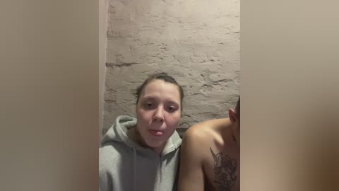 Video of a young woman with light skin and brown hair in a grey hoodie, standing in a brick-walled restroom. She has a tattoo on her right shoulder.