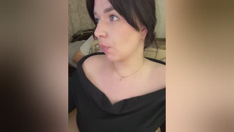 Video of a fair-skinned woman with dark hair, wearing a black off-shoulder top, and a delicate necklace, looking to the left with a neutral expression in a dimly lit room.