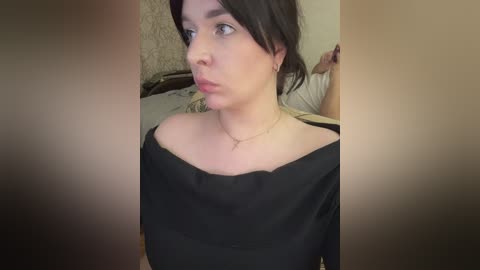 Video of a young woman with fair skin and dark hair, wearing a black off-shoulder top, in a dimly lit room with beige walls and a bed.