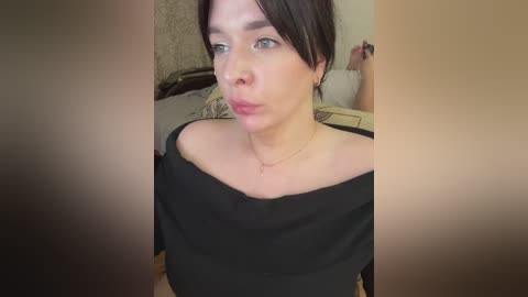 A video of a fair-skinned woman with brown hair, wearing a black off-shoulder top, making a pouty face. Background shows a beige wall and a white bed with pillows.