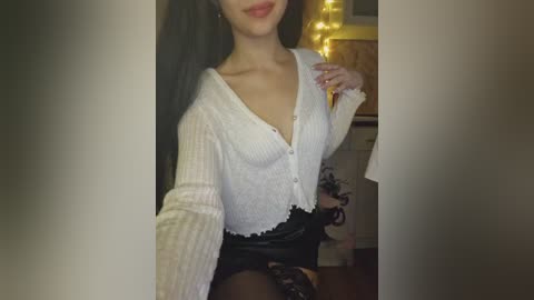 Video of a young woman with long dark hair, wearing a white lace cardigan, black leather skirt, and sheer black stockings, posing indoors near a string of fairy lights.