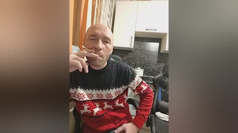 Video of a bald, middle-aged man in a red and black patterned sweater, sitting in a kitchen with white cabinets and a black granite countertop, holding a cigarette to his lips.