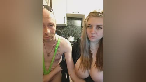Video of a bald, muscular, shirtless man with a green lanyard and a busty, blonde woman in a black jacket, seated in a modern kitchen.
