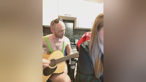 Video of a bald, muscular man with a beard, wearing a neon green tank top, strumming an acoustic guitar in a modern kitchen with white cabinets and dark countertops.
