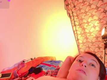 A video of a woman with light skin and dark hair, lying on a colorful, patterned blanket. She has a relaxed expression, with one hand behind her head. The background features a yellow-orange glow and a lace curtain.