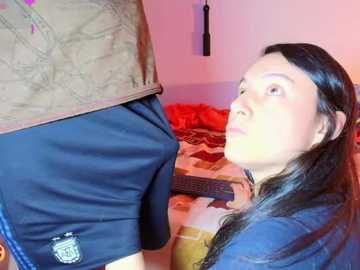 A video shows a young woman with long black hair kneeling before a man in a brown jacket and blue shorts. The room has a red wall, guitar, and black blind hanging, with a casual, intimate atmosphere.