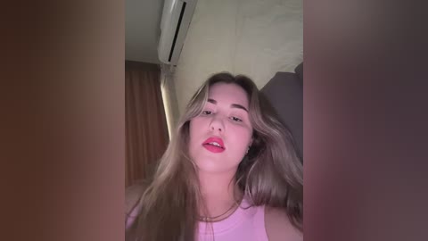 Video of a young woman with long, blonde hair, wearing a pink tank top, leaning forward, looking up, in a dimly lit room with an air conditioning unit and brown curtains in the background.