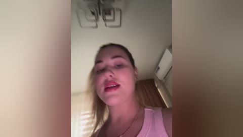 Video of a smiling, light-skinned woman with blonde hair, wearing a pink top, standing indoors against a beige wall, with a ceiling fan and wooden floor visible in the background.