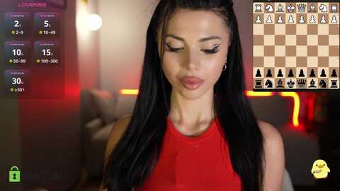 Video of a young woman with long black hair, wearing a red sleeveless top, playing chess on a tablet. Background includes a blurred, modern room with red lighting.