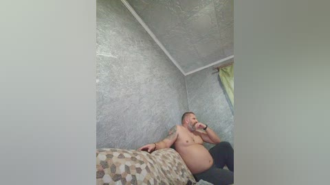A video of a topless, muscular, pregnant man with a beard, lying on a bed in a dimly lit room with textured gray walls and a green curtain.