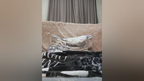 A video of a beige couch with a plush texture, featuring a pillow with a white floral pattern. Clothes, including black and white striped pants, are neatly folded on the couch. The background has brown curtains.