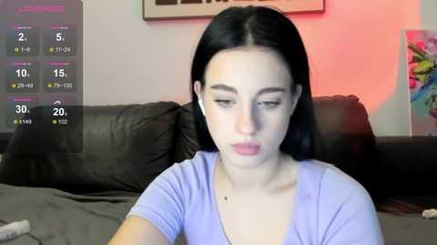 Video of a young woman with fair skin, long dark hair, and a neutral expression, wearing a light purple V-neck top, sitting on a dark couch. Background includes a colorful, abstract painting and a digital display showing weather and temperature.