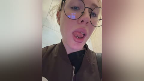 Video of a fair-skinned woman with blonde hair, wearing glasses and a brown jacket, licking her tongue out, in a bathroom with tiled walls.