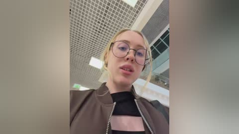 Video of a blonde woman with glasses, wearing a black top and brown jacket, standing in an indoor setting with a grid ceiling and glass windows.