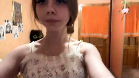 Video of a young woman with light skin, brown hair, wearing a lace tank top, standing in a cozy room with orange curtains, black cat, and framed photos on the wall.