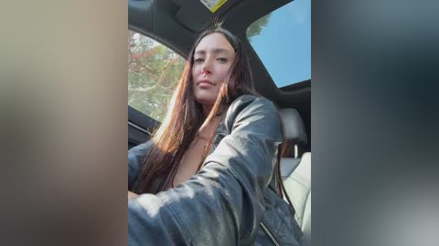 Video of a young woman with long, straight brown hair, fair skin, and medium build, wearing a black leather jacket, sitting in a car with a view of trees outside.