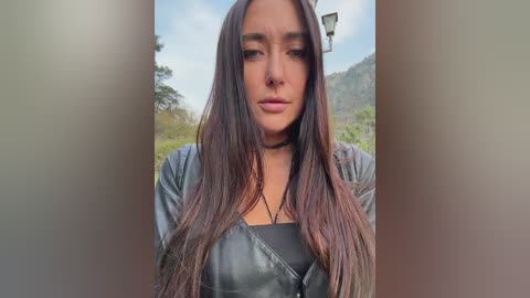 Video of a young woman with long, dark hair, wearing a black leather jacket, standing outdoors in a blurred, green background with a street lamp.