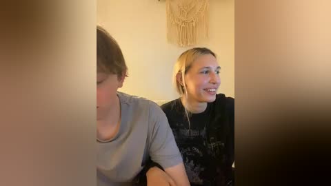 Video of two young women seated closely, one with short hair in a light gray shirt, the other with blonde hair in a black graphic tee, smiling, in a cozy, warmly lit room with a macram\u00e9 wall hanging.