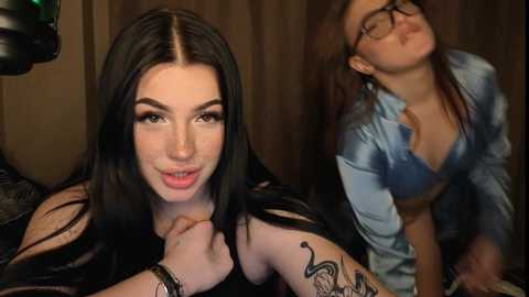 Video of two women: a fair-skinned woman with long black hair, wearing glasses, and a tattooed arm, and a fair-skinned woman with long brown hair, wearing a blue blouse. Both smiling.