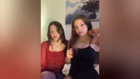 Video of two young women in a dimly lit room. One with long brown hair in a red top, the other with straight brown hair in a black top, both holding drinks.