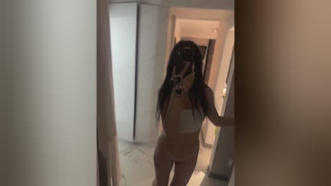 Video of a young woman with long black hair, wearing a white bandeau top and matching thong, taking a mirror selfie in a dimly lit hallway, capturing her slender physique and small breasts.