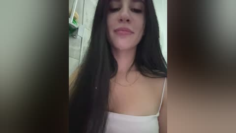 Video of a young woman with long dark hair, wearing a white spaghetti-strap top, smiling slightly, in a dimly lit room with blurred background.