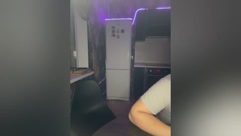 Video of a dimly lit, cramped kitchen with a black chair, white refrigerator, and purple LED strip lights. The scene appears slightly blurred, giving a hazy, somewhat surreal ambiance.