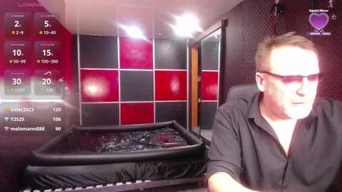 Video of a man in a black shirt, sitting in a dimly lit room with a red and black checkered wall, looking at a black, water-filled bathtub.