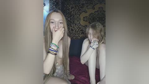 Video of two young women with pale skin, long blonde hair, and small breasts. One is topless, wearing multiple beaded bracelets, sitting on a bed with a red blanket. The other, nude, covers her face with her hands.
