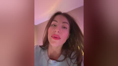 A video of a young Asian woman with long, dark hair, wearing a light blue shirt, smiling slightly. The background is blurred, with a hint of pink lighting.