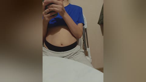 A video of a light-skinned person with a small belly, wearing a blue shirt and grey sweatpants, taking a mirror selfie in a bathroom.