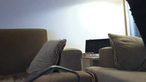 Video of a dimly lit living room with beige couches, a laptop on a table, and a large window partially covered by sheer curtains.