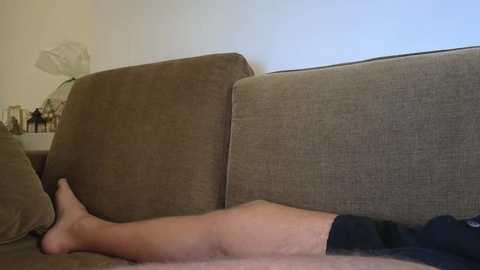 Video of a person lying on a beige couch, wearing black shorts, with bare legs stretched out, against a light-colored wall.