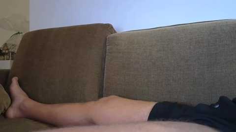 Video of a light-skinned person's bare legs and feet resting on a beige couch against a light blue wall. The person is wearing black shorts.