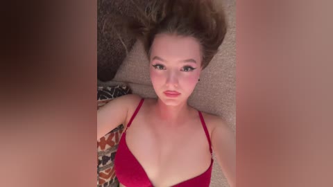 Video of a fair-skinned woman with light brown hair, wearing a red bra, lying on a patterned rug, with a blurred, dark-haired person in the foreground.