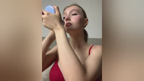 A video of a young Asian woman with fair skin, wearing a red spaghetti-strap top, applying lipstick in front of a mirror, framed by a beige curtain.