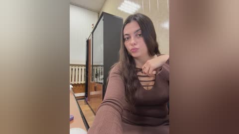 Video of a young woman with long brown hair, wearing a brown, sheer top, sitting on a wooden floor, in a dimly lit room with white walls and a radiator.