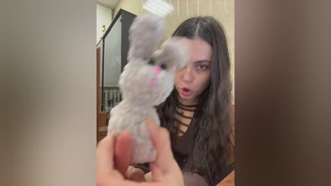 A video shows a woman with long brown hair, dressed in a striped top, holding a fluffy white bunny with pink ears. The background is a dimly lit room with beige walls.