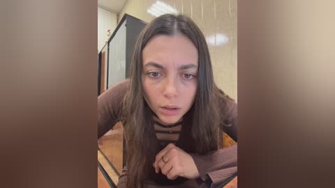 Video of a young woman with long brown hair and fair skin, wearing a striped brown sweater, lying on a wooden floor in a dimly lit room with beige walls.