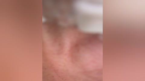 The image is a blurry video of a close-up of a person's genitalia, featuring a penis and testicles. The skin tone is light, and the background is out of focus, with indistinct colors.