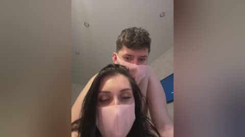 Video of a shirtless young man with short brown hair and a mask, holding a masked woman with long black hair and a mask, both indoors.